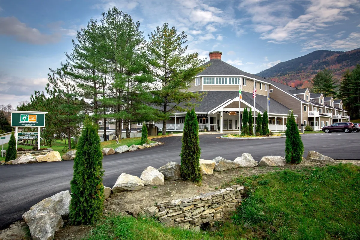 The image captures the main entrance of the hotel, framed by mature trees and landscaped stonework, with flag poles enhancing the sense of arrival. The backdrop reveals glimpses of the surrounding mountains and lush greenery, highlighting the resort’s connection to nature and the outdoors.