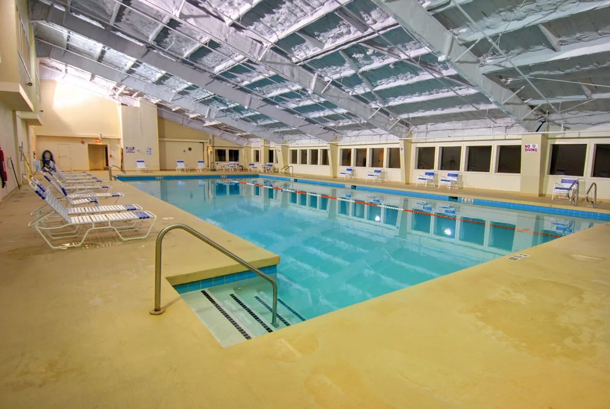This image showcases a spacious indoor swimming pool surrounded by comfortable lounge chairs and illuminated by natural light streaming through large windows. The inviting pool area features an open ceiling structure, creating a bright and relaxing environment ideal for year-round swimming, family fun, and social gatherings.