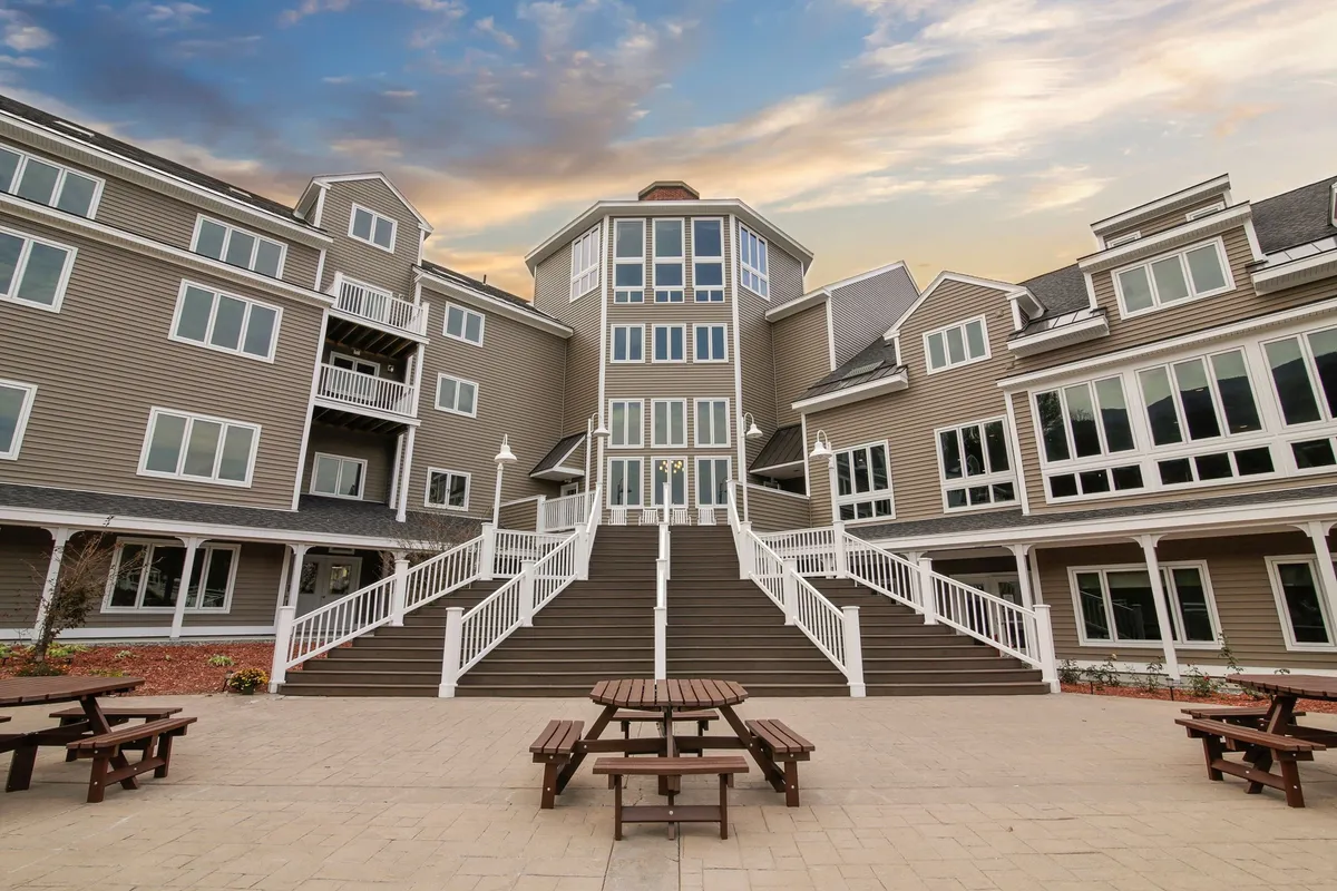 The central entrance with large glass windows and a welcoming staircase is flanked by buildings with balconies. The area features picnic tables where families and friends gather, with the glow of sunset creating an inviting, communal outdoor atmosphere.