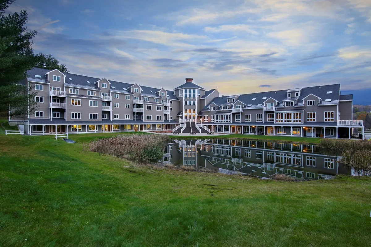 This image showcases the exterior of the property, focusing on the building’s central entrance, a tranquil reflecting pond, and lush surrounding greenery. The scene captures people strolling along a path beside the water and relaxing in view of the sunset.