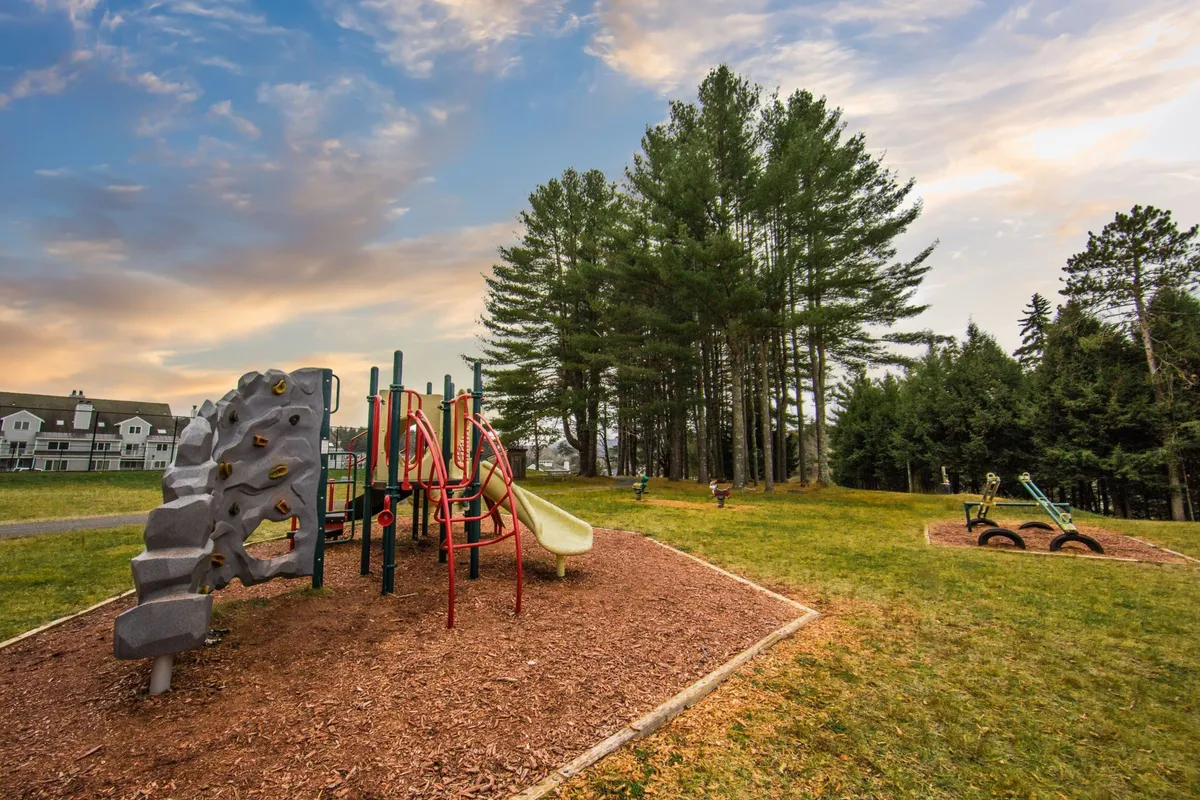The image features an inviting outdoor area with playground equipment nestled among trees, surrounded by wooden mulch. The scene is lively, with children playing, families gathering, and the soft, warm glow of evening sunlight filtering through the trees.