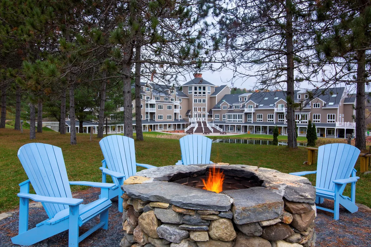 A cozy outdoor fire pit area is surrounded by blue Adirondack chairs, positioned in front of the lodge building. The sun sets in the background, casting a warm glow and inviting guests to gather, relax, and share stories after a day spent outdoors.