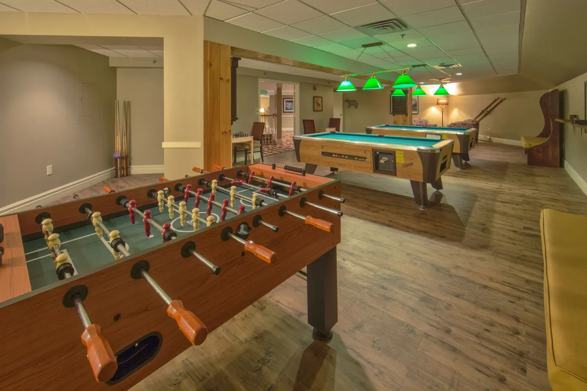 A lively, casual game room featuring foosball and pool tables beneath stylish green pendant lights. The scene suggests friendly competition and relaxed socializing, inviting guests of all ages to gather for memorable moments and shared fun.